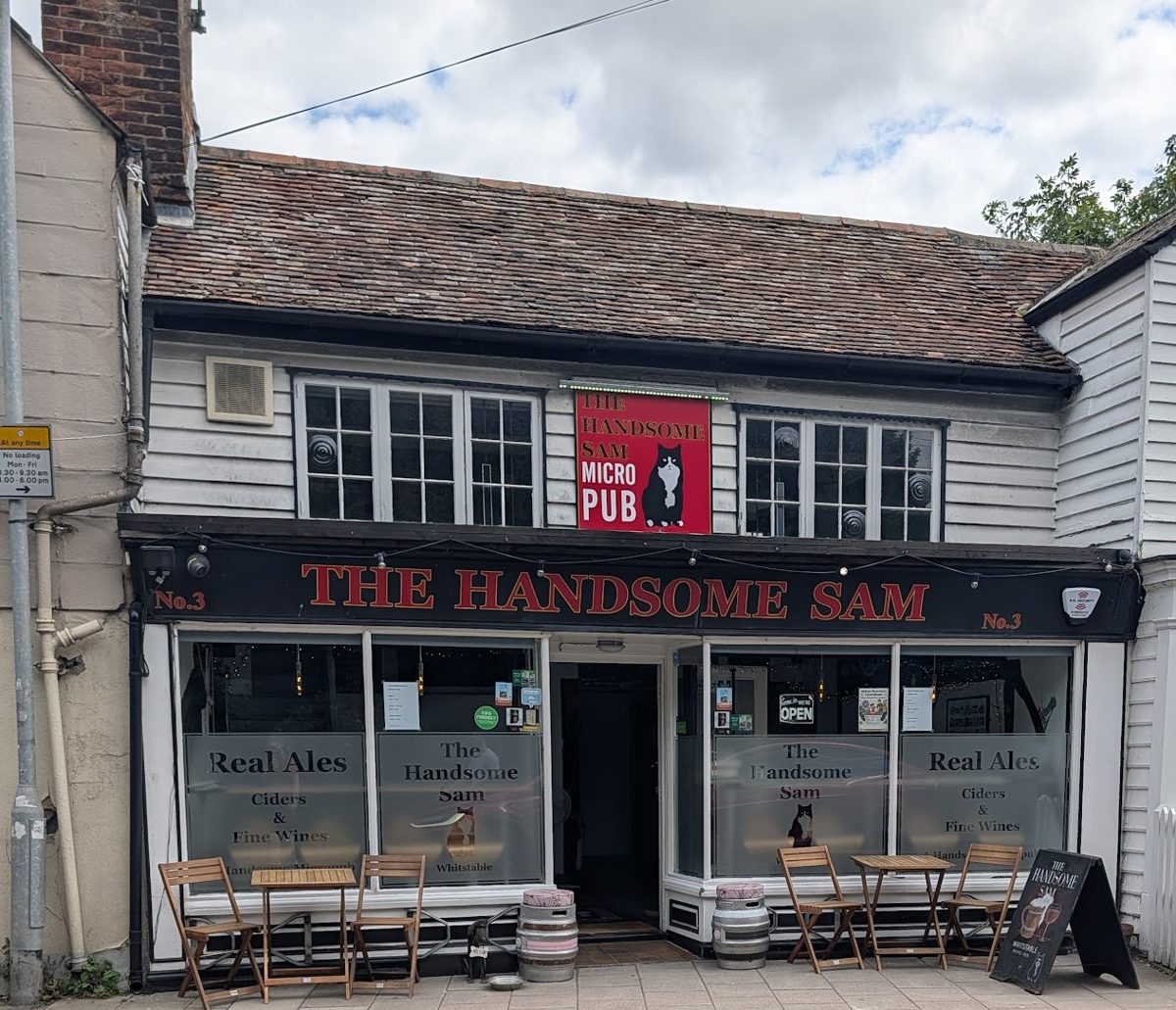 The Handsome Sam front, with a red cat sign, outdoor tables and casks by the door